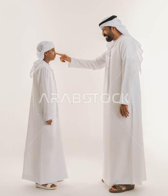 تحمل المسؤولية والاعتناء بالأولاد، الرابطة الأبوية والترابط الأسري، تعابير السعادة والسرور، ايماءات الحب والعطف على الأبناء، بورتريه من الجانب لرجل عربي خليجي اماراتي يرتدي الكندورة والغترة يشير بإصبع السبابة الى وجه ابنه، صورة الجسم بالطول الكامل، خلفية بيضاء