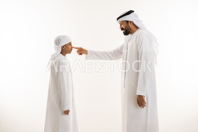 مفهوم الرابطة الأبوية والترابط الأسري، تعابير السعادة والسرور، ايماءات الحب والعطف على الأبناء، تحمل المسؤولية والاعتناء بالأولاد، بورتريه مقرب من الجانب لرجل عربي خليجي اماراتي يرتدي الكندورة والغترة يشير بإصبع السبابة الى وجه ابنه ، خلفية بيضاء