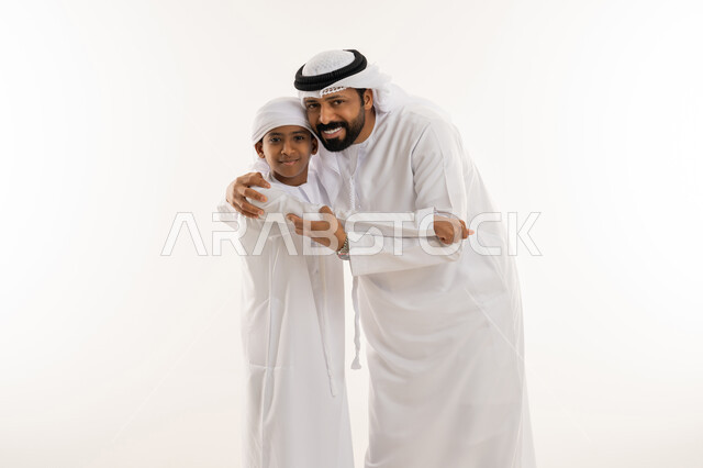 النظر للكاميرا بتعابير السعادة والسرور، مفهوم الرابطة الأبوية والترابط الأسري، ايماءات الحب والعطف على الأبناء، تحمل المسؤولية والاعتناء بالأولاد، بورتريه مقرب لرجل عربي خليجي اماراتي يرتدي الكندورة والغترة يحتضن ابنه، خلفية بيضاء