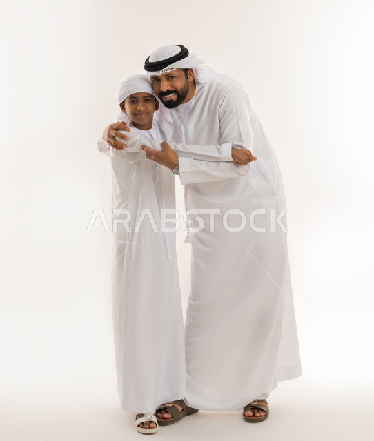 ايماءات الحب والعطف على الأبناء، تحمل المسؤولية والاعتناء بالأولاد، مفهوم الرابطة الأبوية والترابط الأسري، بورتريه لرجل عربي خليجي اماراتي يرتدي الكندورة والغترة يحتضن ابنه، النظر للكاميرا بتعابير السعادة والسرور، صورة الجسم بالطول الكامل، خلفية بيضاء