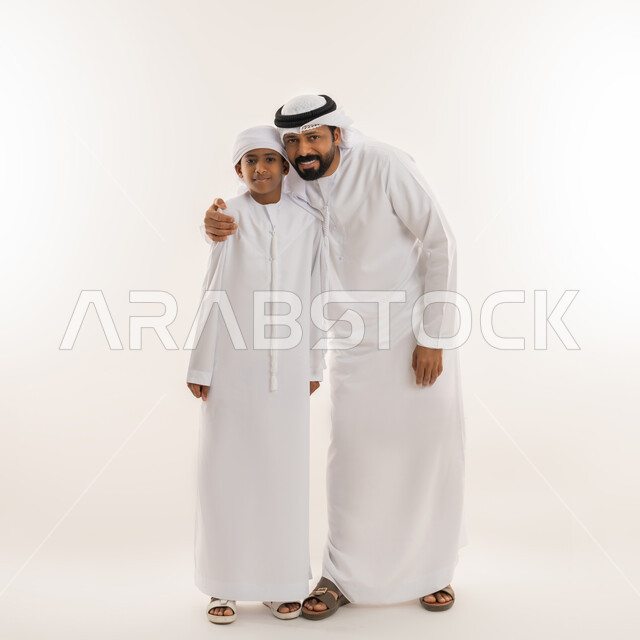 تحمل المسؤولية والاعتناء بالأولاد، مفهوم الرابطة الأبوية والترابط الأسري، ايماءات الحب والعطف على الأبناء، بورتريه لرجل عربي خليجي اماراتي يرتدي الكندورة والغترة يحتضن ابنه، النظر للكاميرا بتعابير السعادة والسرور، صورة الجسم بالطول الكامل، خلفية بيضاء