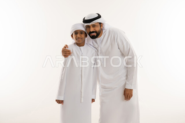 مفهوم الرابطة الأبوية والترابط الأسري، ايماءات الحب والعطف على الأبناء، تحمل المسؤولية والاعتناء بالأولاد، بورتريه مقرب لرجل عربي خليجي اماراتي يرتدي الكندورة والغترة يحتضن ابنه، النظر للكاميرا بتعابير السعادة والسرور، خلفية بيضاء