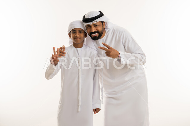 تعابير السعادة بالوصول للغايات، مفهوم الفوز والانتصار، النجاح وتحقيق الأهداف، الألفة والمحبة بين الأفراد، بورتريه مقرب لرجل عربي خليجي اماراتي يرتدي الكندورة والغترة يقف برفقة ابنه يرفعون ايديهم بإشارة النصر والقوة، النظر للكاميرا بإيماءات الفرح، خلفية بيضاء