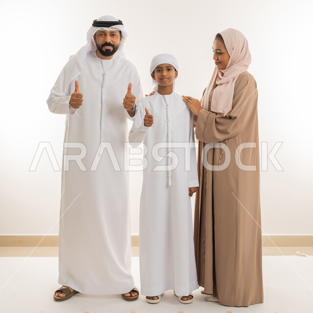 رعاية الآباء بأبنائهم والاهتمام بهم، تعابير الإعجاب والقبول، الترابط الأسري والحفاظ على الاستقرار العائلي، بورتريه لعائلة عربية اماراتية يرفعوا اصبع الابهام للأعلى بهدف الرضا والموافقة، النظر للكاميرا بإيماءات السعادة، صورة الجسم بالطول الكامل، خلفية بيضاء