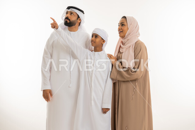 مفهوم لغة الجسد، النظر الى مكان معين، التوجه والإشارة الى شيء ما، رعاية لآباء بالأبناء والاهتمام بهم، بورتريه مقرب لولد عربي خليجي اماراتي رتدي الكندورة والعمامة يقف برفقة والده و والدته يشير بإصبع السبابة الى الأعلى دلالة على موقع محدد، خلفية بيضاء