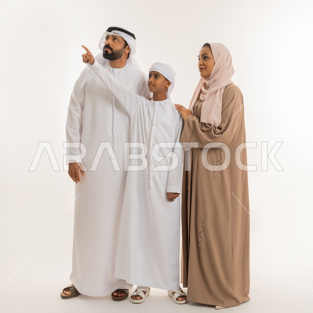 التوجه والإشارة  لشيء ما، رعاية لآباء بالأبناء والاهتمام بهم، النظر الى مكان معين، مفهوم لغة الجسد، بورتريه لولد عربي خليجي اماراتي رتدي الكندورة والعمامة يقف برفقة عائلته يشير بإصبع السبابة الى الأعلى دلالة على موقع محدد، صورة الجسم بالطول الكامل، خلفية بيضاء