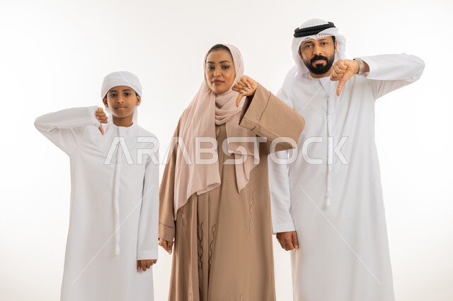 مفهوم التقييم السلبي والرفض، الشعور بالإنزعاج من شيء ما، بورتريه مقرب لرجل عربي خليجي امارتي يرتدي الكندورة والغتره يقف برفقة زوجته وابنه يشيروا بإصبع الابهام الى الاسفل دلالة على عدم الموافقة، النظر للكاميرا بتعابير الفشل، خلفية بيضاء