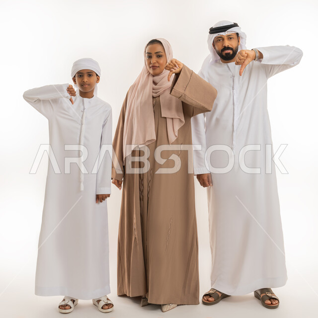Feeling upset about something, concept of negative evaluation and rejection, portrait of an Arab Gulf Emirati man wearing a kandura and ghutra standing with his wife and son pointing thumbs down as a sign of disapproval, looking at the camera with an expression of failure, full body image, white background