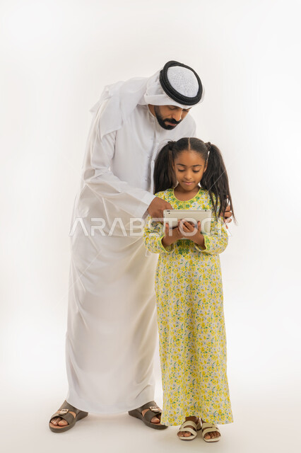 مشاهدة الأنشطة الترفيهية والتعليمية، اهتمام الآباء بالأبناء، دمج التعلم بالتكنولوجيا، متابعة منصات التعليم الالكتروني عبر الانترنت، بورتريه لفتاة عربية خليجية اماراتية ترتدي لباس الكاجوال تستخدم التابلت وتقف برفقة والدها، صورة الجسم بالطول الكامل، خلفية بيضاء