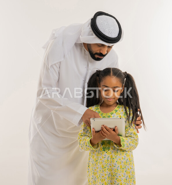 دمج التعلم بالتقنية والتكنولوجيا، متابعة منصات التعليم الالكتروني عبر الانترنت، اهتمام الآباء بالأبناء، مشاهدة الأنشطة الترفيهية والتعليمية، بورتريه مقرب لفتاة عربية خليجية اماراتية ترتدي لباس الكاجوال تستخدم التابلت وتقف برفقة والدها، خلفية بيضاء