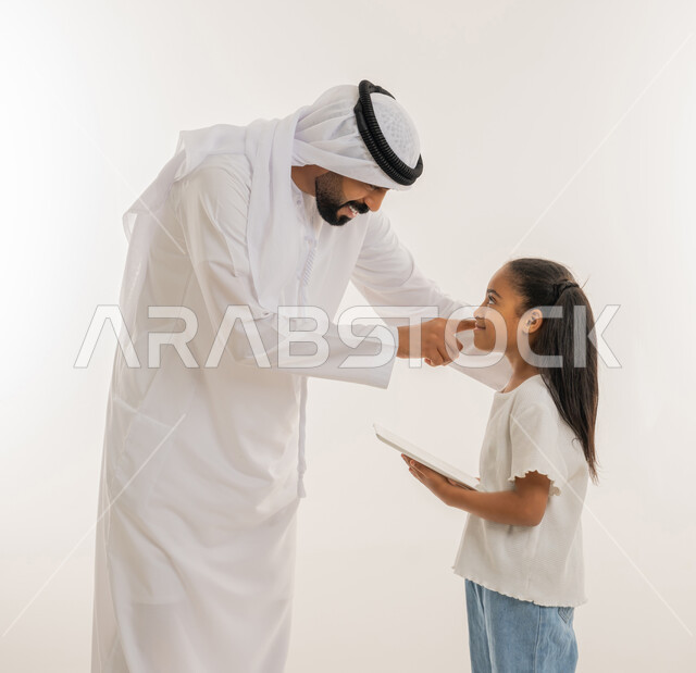 تعابير السعادة والسرور، متابعة منصات التعليم الالكتروني المتاحة عبر الانترنت، دمج التعلم مع التقنية والتكنولوجيا، مشاهدة الأنشطة الترفيهية والتعليمية، بورتريه مقرب لفتاة عربية خليجية اماراتية ترتدي لباس الكاجوال تقف برفقة والدها وتستخدم التابلت، خلفية بيضاء