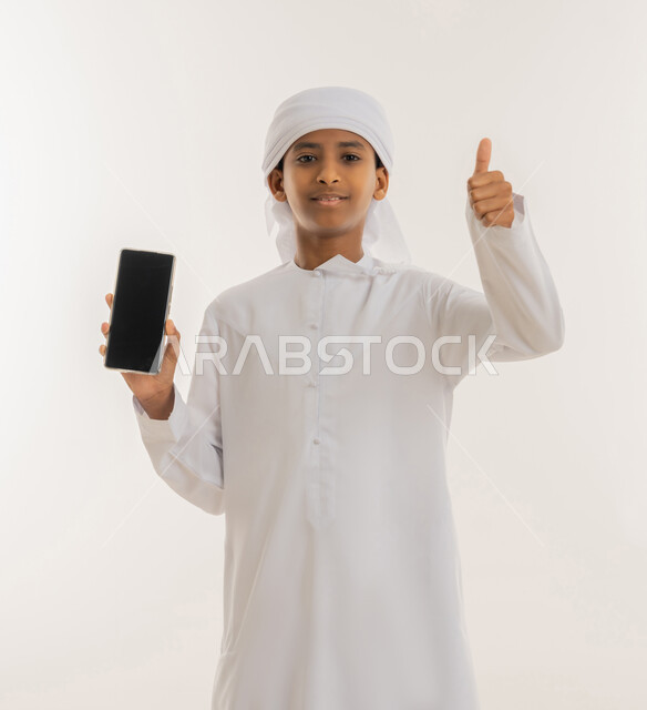 Using modern and advanced technology and techniques, raising the thumb up with the aim of admiration and acceptance, looking at the camera with gestures of happiness and pleasure, close-up portrait of an Arab Gulf Emirati boy wearing a kandura and turban using a mobile phone, displaying a blank black screen, white background