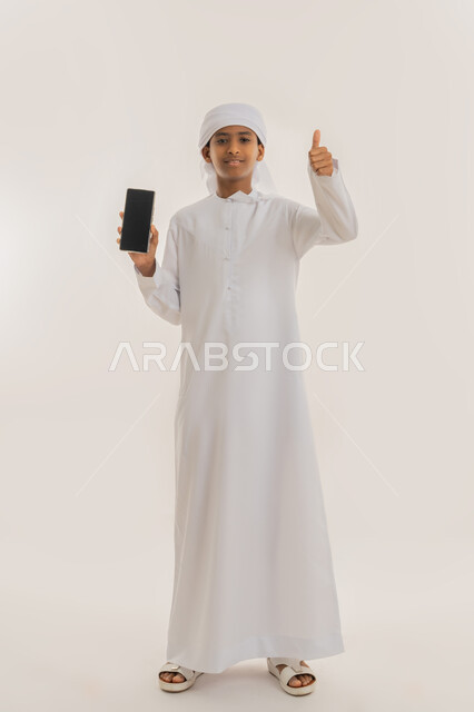 Thumbs up showing approval, using advanced technology, looking at camera with gestures of happiness and pleasure, portrait of an Arab Gulf Emirati boy wearing a kandura and turban using a mobile phone, displaying a blank black screen, full-length body image, white background