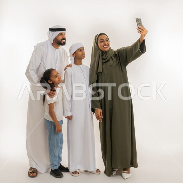 أجواء عائلية سعيدة مليئة بالحب، الألفة والمحبة بين أفراد الأسرة، التقاط صور تذكارية، استخدام التقنية الحديثة، بورتريه لإمرأة عربية خليجية اماراتية ترتدي الحجاب والعباءة تقف برفقة عائلتها تستخدم الهاتف بتعابير السعادة، صورة الجسم بالطول الكامل، خلفية بيضاء
