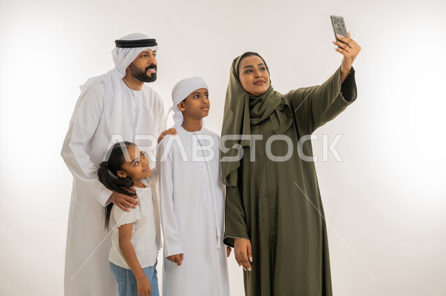 الألفة والمحبة بين أفراد الأسرة، التقاط صور تذكارية، أجواء عائلية سعيدة مليئة بالحب، استخدام التقنية الحديثة، بورتريه مقرب لإمرأة عربية خليجية اماراتية ترتدي الحجاب والعباءة تقف برفقة عائلتها تستخدم الهاتف بتعابير السعادة والسرور، خلفية بيضاء