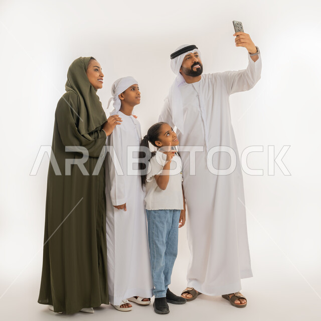 التقاط صور تذكارية، الألفة والمحبة بين أفراد الأسرة، أجواء عائلية سعيدة مليئة بالحب، استخدام التقنية الحديثة، بورتريه لرجل عربي خليجي اماراتي يرتدي الكندورة والغترة يقف برفقة عائلته يستخدم الهاتف بتعابير السعادة والسرور، صورة الجسم بالطول الكامل، خلفية بيضاء