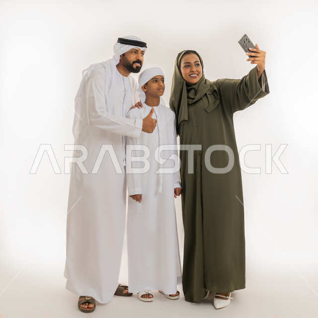 استخدام التقنية الحديثة، الألفة والمحبة بين أفراد الأسرة، أجواء عائلية سعيدة مليئة بالحب، التقاط صور تذكارية، بورتريه لإمرأة عربية خليجية اماراتية ترتدي الحجاب والعباءة تقف برفقة زوجها وابنها تستخدم الهاتف بتعابير السعادة، صورة الجسم بالطول الكامل، خلفية بيضاء