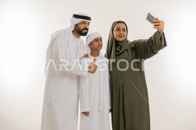 أجواء عائلية سعيدة مليئة بالحب، الألفة والمحبة بين أفراد الأسرة، استخدام التقنية الحديثة، التقاط صور تذكارية، بورتريه مقرب لإمرأة عربية خليجية اماراتية ترتدي الحجاب والعباءة تقف برفقة زوجها وابنها تستخدم الهاتف المحمول بتعابير السعادة والسرور، خلفية بيضاء