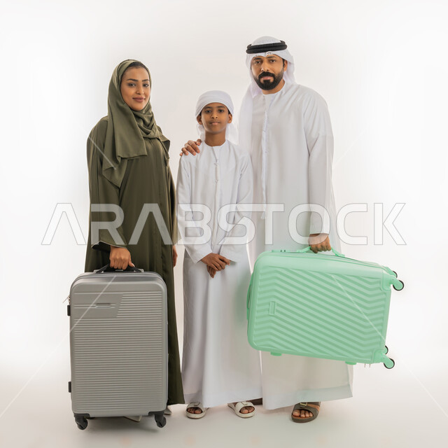 النظر الى الكاميرا بإيماءات السعادة والسرور، الاستعداد للسفر في جولة سياحية، مفهوم السياحة في إجازة الصيف، بورتريه مقرب لرجل عربي خليجي اماراتي يرتدي الكندورة والغترة يقف برفقة زوجته وابنه مبتسمين ويمسكون حقائب الأمتعة، صورة الجسم بالطول الكامل، خلفية بيضاء