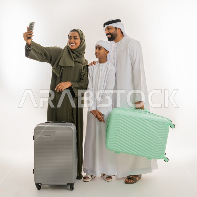 مفهوم السياحة في إجازة الصيف، الاستعداد للسفر في جولة سياحية، استخدام التقنية الحديثة، بورتريه لإمرأة عربية خليجية اماراتية ترتدي الحجاب والعباءة تقف برفقة ابنها وزوجها وتستخدم الهاتف المحمول، رجل يمسك بيده حقيبة الأمتعة، صورة الجسم بالطول الكامل، خلفية بيضاء