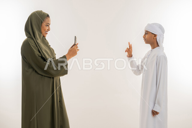 مفهوم السعادة بالوصول للغايات، استخدام التقنية الحديثة، النجاح وتحقيق الأهداف، بورتريه مقرب من الجانب لإمرأة عربية خليجية اماراتية ترتدي الحجاب والعباءة تستخدم الهاتف، ولد يرتدي الكندورة والعمامة يرفع يده بإشارة النصر والقوة، خلفية بيضاء