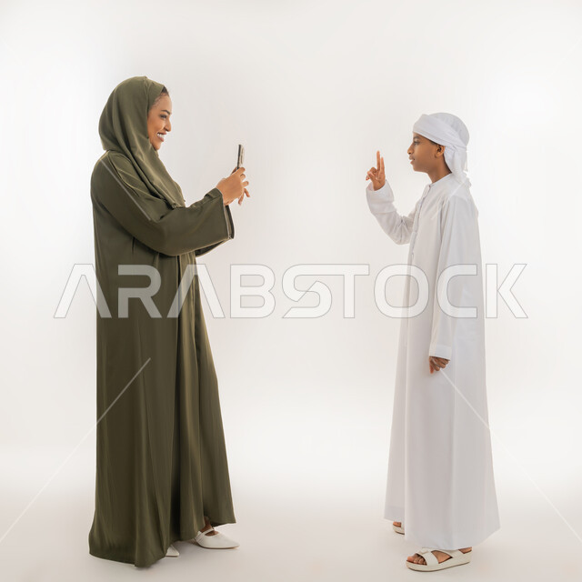 استخدام التقنية الحديثة، النجاح وتحقيق الأهداف، مفهوم السعادة بالوصول للغايات، بورتريه من الجانب لإمرأة عربية خليجية اماراتية ترتدي الحجاب والعباءة تستخدم الهاتف، ولد يرتدي الكندورة والعمامة يرفع يده بإشارة النصر والقوة، صورة الجسم بالطول الكامل، خلفية بيضاء