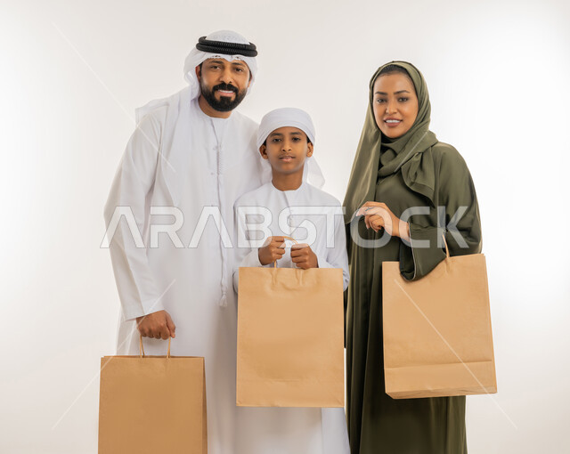 النظر الى الكاميرا بإيماءات السعادة بعد التسوق والشراء وتأثيره الايجابي، عروضات وتخفيضات موسمية في الأسواق، بورتريه مقرب لرجل عربي خليجي اماراتي يرتدي الكندورة والغترة يقف برفقة زوجته وابنه ويحملون الأكياس الورقية، خلفية بيضاء