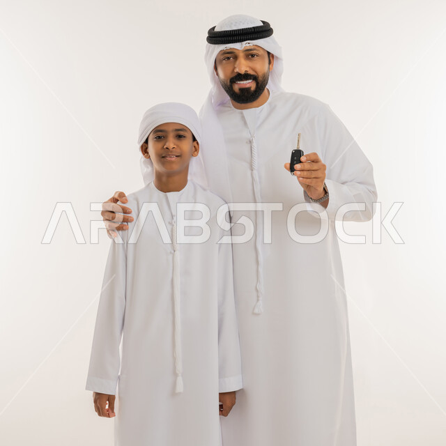 النظر الى الكاميرا بإيماءات السعادة والسرور، فرحة تملك شيء جديد، الحصول على رخصة القيادة، امتلاك سيارة جديدة، بورتريه مقرب لرجل عربي خليجي اماراتي يرتدي الكندورة والغترة يحمل بيده جهاز التحكم الالكتروني يقوم بمفاجأة ابنه، خلفية بيضاء