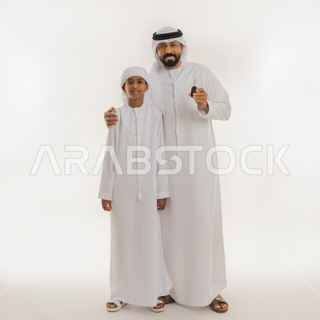Joy of owning something new, getting a driver's license, owning a new car, looking at the camera with gestures of happiness and pleasure, portrait of an Arab Gulf Emirati man wearing a kandura and ghutra holding an electronic control device in his hand surprising his son, full body image, white background