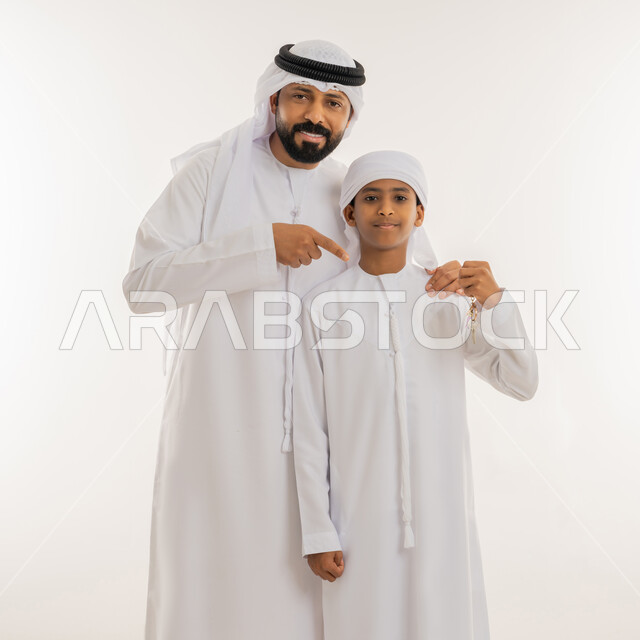 الشعور بالفرح لامتلاك شقة سكنية جديدة، النظر الى الكاميرا بإيماءات السعادة، بورتريه مقرب لولد عربي خليجي اماراتي يرتدي الكندورة والعمامة يقف بجانب والده ويحمل مفتاح المنزل، رجل يشير بإصبع السبابة الى المفاتيح بتعابير السرور، خلفية بيضاء
