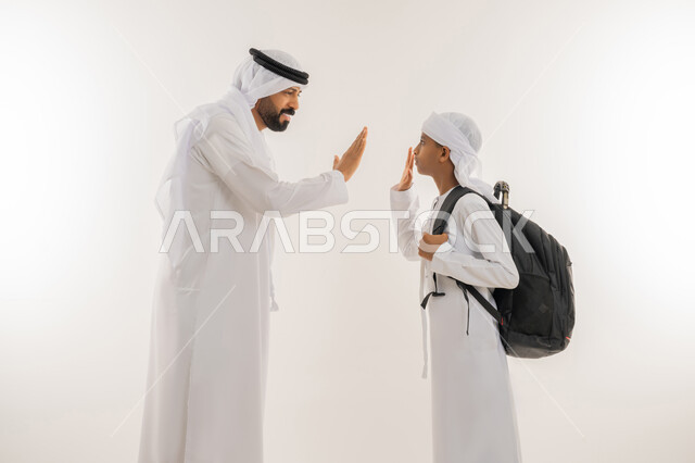 الشعور بالأمان والحب مع الآباء، موسم العودة للمدارس، مفهوم الاعتناء بالأبناء والاهتمام بهم، بورتريه مقرب من الجانب لولد عربي خليجي اماراتي يرتدي الكندورة والعمامة يضع الحقيبة على ظهره يقف برفقة والده ويرفعون ايديهم الى الاعلى بهدف التحية والسلام، خلفية بيضاء