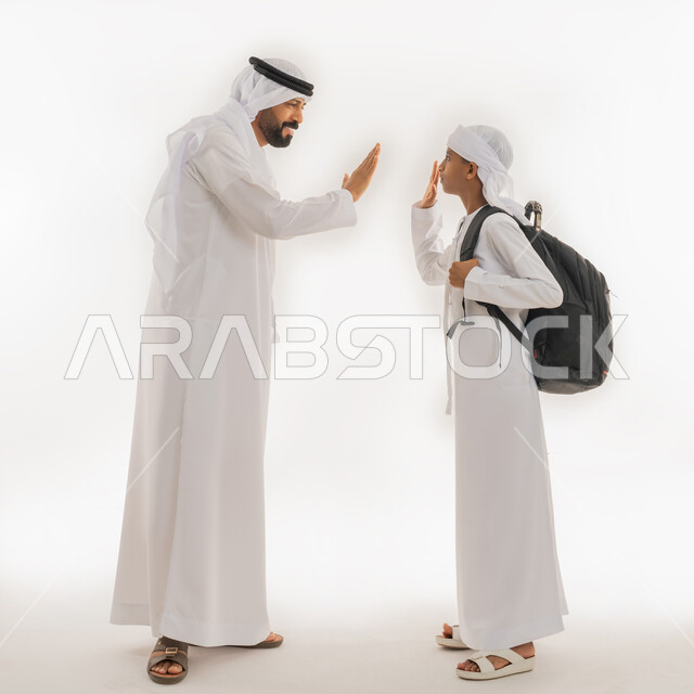 Back to school season, concept of caring for children, feeling safe and loved with parents, portrait of an Arab Gulf Emirati boy wearing a kandura and turban, putting a bag on his back, standing with his father and raising their hands up in greeting, full body image, white background