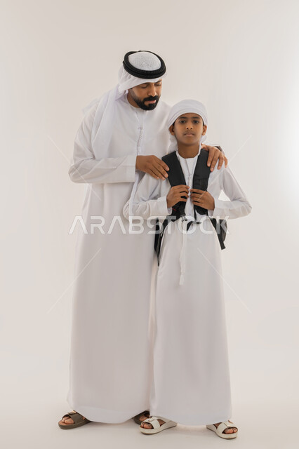 مفهوم الاعتناء بالأبناء والاهتمام بهم، الشعور بالأمان والحب مع الآباء، موسم العودة للمدارس، بورتريه لولد عربي خليجي اماراتي يرتدي الكندورة والعمامة يضع الحقيبة على ظهره ويقف برفقة والده، النظر للكاميرا بتعابير السعادة، صورة الجسم بالطول الكامل، خلفية بيضاء