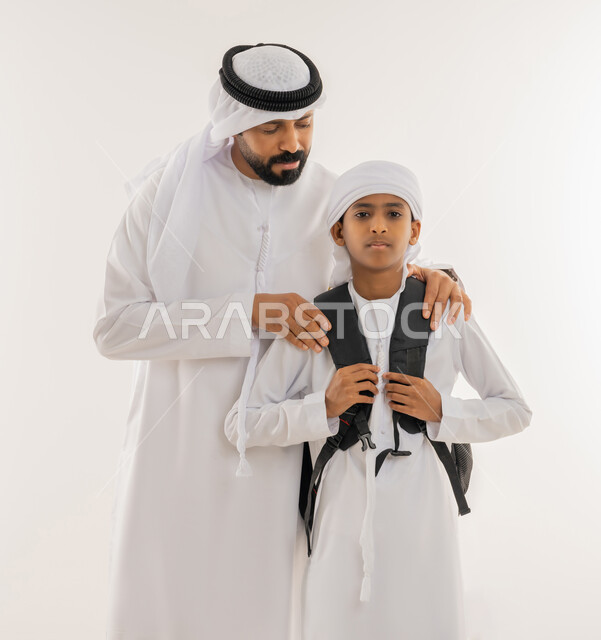 الشعور بالأمان والحب مع الآباء، موسم العودة للمدارس في الامارات، مفهوم الاعتناء بالأبناء والاهتمام بهم، بورتريه مقرب لولد عربي خليجي اماراتي يرتدي الكندورة والعمامة يضع الحقيبة على ظهره ويقف برفقة والده، النظر الى الكاميرا بتعابير السعادة والسرور، خلفية بيضاء