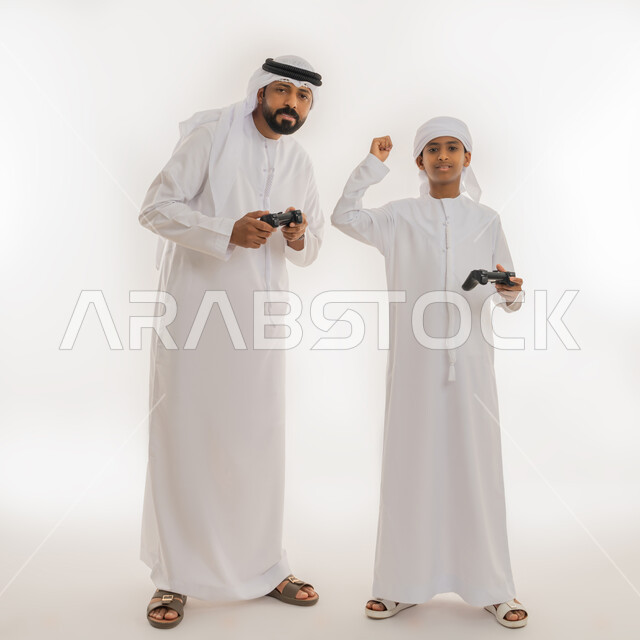 الاستمتاع بالتقنية الحديثة، النظر للكاميرا بتعابير السعادة، برامج الكمبيوتر الترفيهية، بورتريه لولد عربي خليجي اماراتي يرتدي الكندورة والعمامة يقف برفقة والده يحملون مقود البلايستيشن ويرفع قبضة يده للأعلى بهدف الحماس والفوز، صورة الجسم بالطول الكامل، خلفية بيضاء