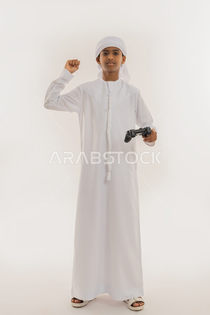 النظر للكاميرا بتعابير السعادة، الاستمتاع بالتكنولوجيا الحديثة، برامج الكمبيوتر الترفيهية، بورتريه لولد عربي خليجي اماراتي يرتدي الكندورة والعمامة يحمل مقود البلايستيشن ويرفع قبضة يده للأعلى بهدف الحماس والفوز، صورة الجسم بالطول الكامل، خلفية بيضاء