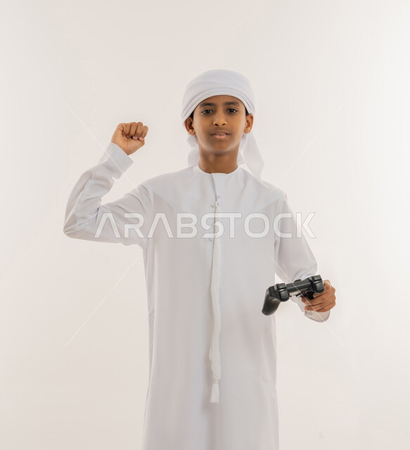 مفهوم المتعة والتسلية، الاستمتاع بالتكنولوجيا الحديثة، برامج الكمبيوتر الترفيهية، بورتريه مقرب لولد عربي خليجي اماراتي يرتدي الكندورة والعمامة يحمل مقود البلايستيشن ويرفع قبضة يده للأعلى بهدف الحماس والفوز، النظر للكاميرا بتعابير السعادة والسرور، خلفية بيضاء