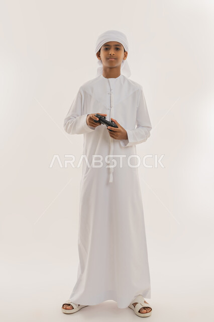 برامج الكمبيوتر الترفيهية، الاستمتاع بالتكنولوجيا والتقنية الحديثة، مفهوم المتعة والتسلية، قضاء وقت ممتع باللعب، بورتريه لولد عربي خليجي اماراتي يرتدي الكندورة والعمامة يحمل قبضة البلايستيشن، النظر للكاميرا بتعابير السعادة، صورة الجسم بالطول الكامل، خلفية بيضاء