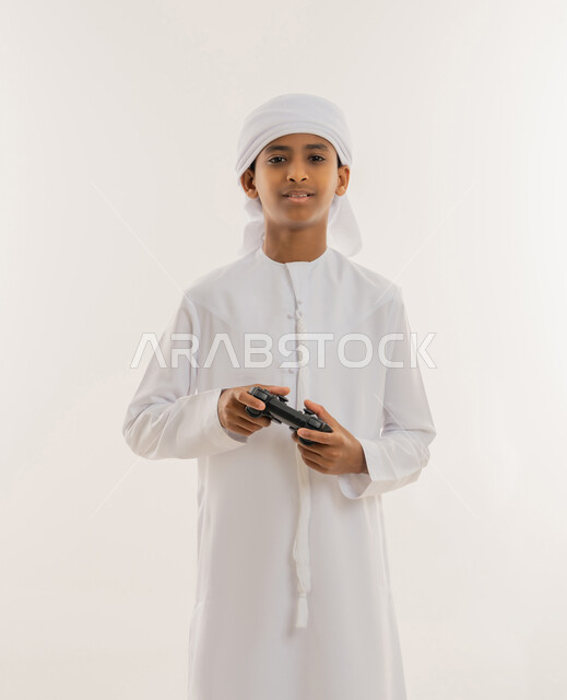 الاستمتاع بالتقنيات والتكنولوجيا الحديثة، برامج الكمبيوتر الترفيهية، مفهوم المتعة والتسلية، قضاء وقت ممتع باللعب، بورتريه مقرب لولد عربي خليجي اماراتي يرتدي الكندورة والعمامة يحمل قبضة البلايستيشن، النظر للكاميرا بتعابير السعادة والسرور، خلفية بيضاء