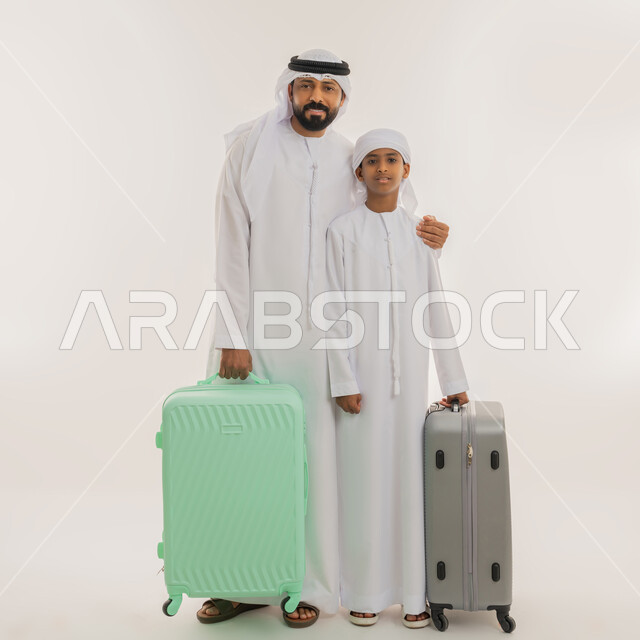 قضاء رحلة صيفية ممتعة، مفهوم السياحة في إجازة الصيف، الاستعداد للسفر في جولة سياحية، بورتريه لأب عربي خليجي اماراتي يرتدي الكندورة والغترة يقف برفقة ابنه ويمسكون بأيديهم حقيبة الأمتعة، النظر الى الكاميرا بإيماءات السعادة، صورة الجسم بالطول الكامل، خلفية بيضاء