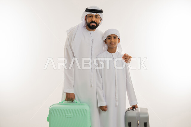 الاستعداد للسفر في جولة سياحية، مفهوم السياحة في إجازة الصيف، قضاء رحلة صيفية ممتعة، بورتريه مقرب لأب عربي خليجي اماراتي يرتدي الكندورة والغترة يقف برفقة ابنه ويمسكون بأيديهم حقيبة الأمتعة، النظر الى الكاميرا بإيماءات السعادة والسرور، خلفية بيضاء
