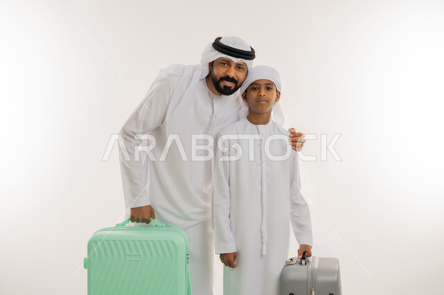 Summer vacation tourism concept, having a fun summer trip, getting ready to travel on a tour, close-up portrait of an Emirati Gulf Arab father wearing a kandura and a ghutra standing with his son and holding a luggage bag in their hands, looking at the camera with gestures of happiness and pleasure, white background