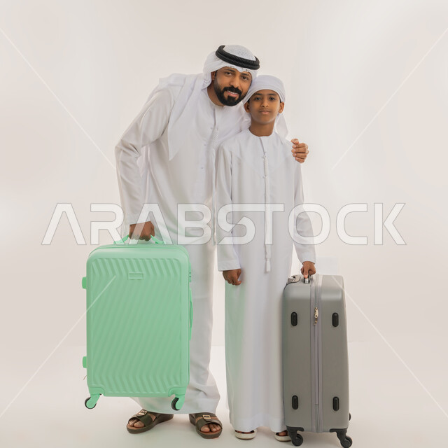 قضاء رحلة صيفية ممتعة، مفهوم السياحة في إجازة الصيف، الاستعداد للسفر في جولة سياحية، بورتريه لأب عربي خليجي اماراتي يرتدي الكندورة والغترة يقف برفقة ابنه ويمسكون بأيديهم حقيبة الأمتعة، النظر الى الكاميرا بإيماءات السعادة، صورة الجسم بالطول الكامل، خلفية بيضاء