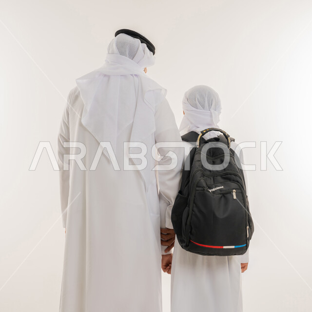 الشعور بالأمان والحب مع الآباء، موسم العودة للمدارس في الامارات، مفهوم الاعتناء بالأبناء والاهتمام بهم، بورتريه مقرب من الخلف لولد عربي خليجي اماراتي يرتدي الكندورة والعمامة يضع الحقيبة على ظهره ويمسك بيد والده، الاستعداد للعام الدراسي، خلفية بيضاء