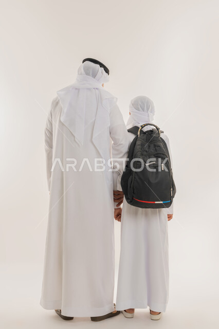 موسم العودة للمدارس في الامارات العربية المتحدة، الشعور بالأمان والحب مع الآباء، مفهوم الاعتناء بالأبناء والاهتمام بهم، بورتريه من الخلف لولد عربي خليجي اماراتي يرتدي الكندورة والعمامة يضع الحقيبة على ظهره ويمسك بيد والده، صورة الجسم بالطول الكامل، خلفية بيضاء