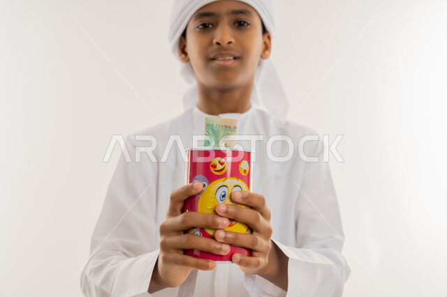 النظر الى الكاميرا بتعابير السعادة والسرور، تربية الأطفال على التوفير وجمع المال، مفهوم الاستثمار المالي وإدخار الأموال ووعي الأولاد، بورتريه مقرب لولد عربي خليجي اماراتي يرتدي الكندورة والعمامة يقوم بتجميع مصروفه داخل الحصالة، خلفية بيضاء