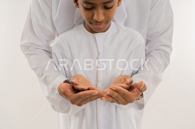 طلب المساعدة من المولى عز وجل، إيماءات التوسل والاسترحام، الحمد والشكر والتضرع  لله تعالى، بورتريه مقرب لولد عربي خليجي اماراتي يرتدي الكندورة يقف امام والده ويقوموا برفع أيديهم للأعلى بهدف الدعاء بتعابير السعادة والرضا، خلفية بيضاء