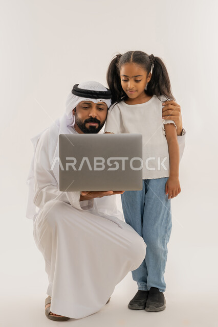 قضاء أوقات ممتعة، التعلم عن بعد وتلقي الدروس عبر الأونلاين، استخدام التكنولوجيا والتقنية الحديثة والمتطورة، أنشطة ترفيهية الكترونية مسلية، بورتريه لرجل عربي خليجي اماراتي يرتدي الكندورة والغترة يقف برفقة ابنته ويستخدم جهاز الحاسوب المحمول، خلفية بيضاء