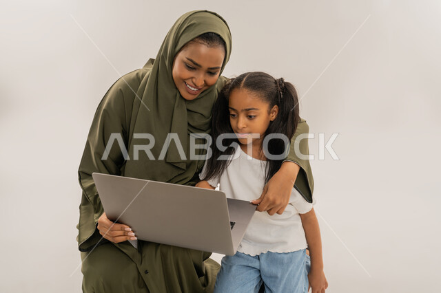 أنشطة ترفيهية الكترونية مسلية، التعلم عن بعد وتلقي الدروس عبر الأونلاين، استخدام التكنولوجيا والتقنية الحديثة والمتطورة، قضاء أوقات ممتعة، بورتريه مقرب لإمرأة عربية خليجية اماراتية ترتدي الحجاب والعباءة تقف برفقة ابنتها وتستخدم جهاز الحاسوب المحمول، خلفية بيضاء