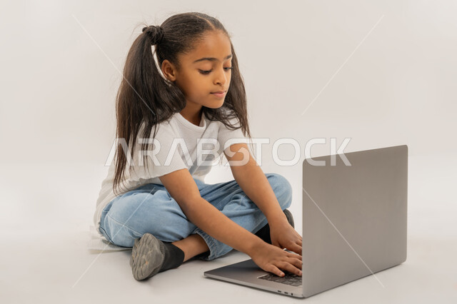 التعلم عن بعد وتلقي الدروس عبر الأونلاين، استخدام التكنولوجيا والتقنية الحديثة والمتطورة، أنشطة ترفيهية الكترونية مسلية، قضاء أوقات ممتعة، بورتريه لفتاة عربية خليجية اماراتية ترتدي لباس الكاجوال تجلس على الأرض وتستخدم جهاز الحاسوب المحمول، خلفية بيضاء